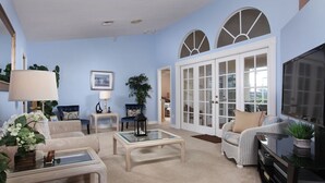 A living room with a beige sofa, a glass coffee table, and a television on a black stand. There are two chairs, a lamp, and a potted plant. - Avail August 28 & on. Sunny New heated Pool & Spa 10min walk2beach 6-bikes (Marco Island)