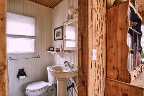 Une salle de bain avec des toilettes, un lavabo et un miroir. Il y a une fenêtre au-dessus des toilettes et une étagère avec des articles de toilette au-dessus du lavabo.