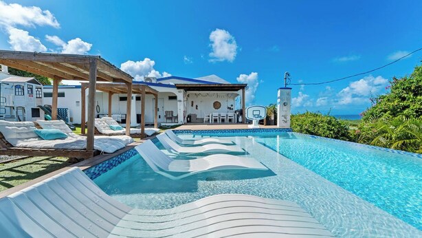 Une piscine avec des chaises longues et une pergola, un bâtiment avec un toit bleu et un ciel d’un bleu limpide.