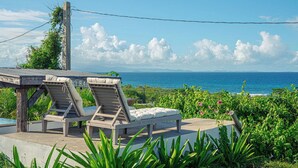 Une terrasse en bois avec deux chaises longues et une petite table, d’où l’on peut admirer une mer d’un bleu limpide et un paysage verdoyant.