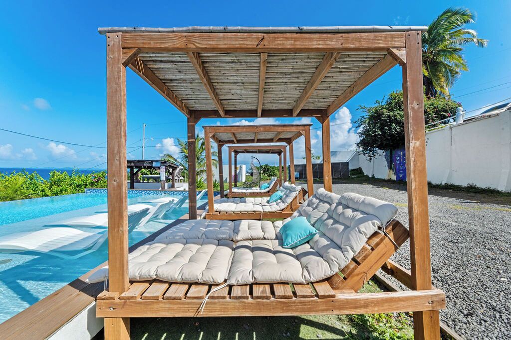A wooden outdoor lounge area with cushioned seating and a pergola overhead, overlooking a swimming pool.