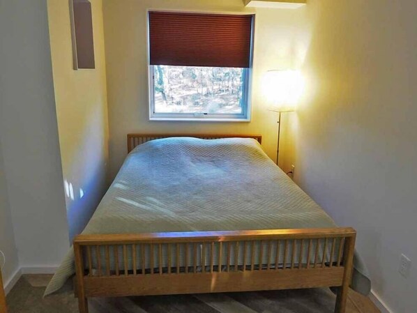 Une petite chambre avec un lit simple, une tête de lit en bois, une fenêtre avec des stores et une lampe de chevet.