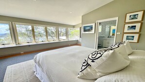 Ein Schlafzimmer mit einem großen Bett, Blick auf Bäume durch das Fenster und einem durchgehenden Türrahmen einsehbaren Badezimmer.