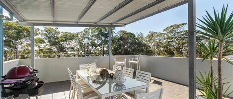 A covered outdoor patio with a table and chairs, a barbecue grill, and potted plants.