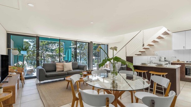 A modern living room with a glass dining table, grey sofas, and a kitchen with white cabinets and a staircase.
