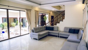 A spacious living room with a large sectional sofa, a staircase, and a sliding glass door leading to an outdoor area with chairs and plants. - 8bdr Avoca Pool Villas 172 (Muang Pattaya)