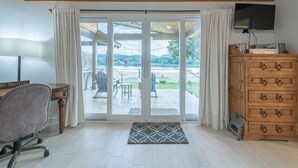 A room with a wooden ceiling, a desk with a chair, a TV on a wooden cabinet, and a sliding glass door leading to an outdoor patio with chairs and a table. - Packsaddle Paradise- Lakefront w/ Beach & Views (Kingsland)