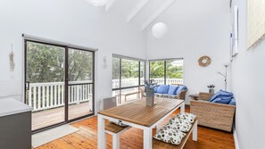 A spacious room with a wooden table, a bench, and a sofa with blue cushions. There are large windows with a view of greenery outside.