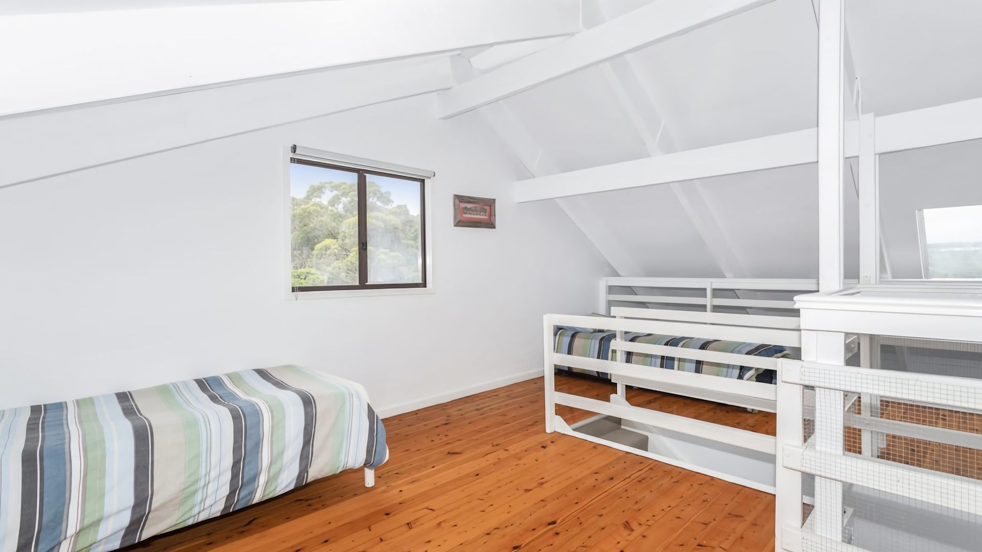 Una habitación con techo inclinado, una cama litera, una cama individual, una ventana con vistas a árboles y un cuadro en la pared.