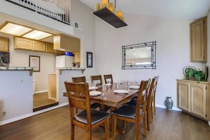 A dining area with a wooden table and chairs, a kitchen in the background, and a mirror on the wall.