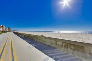 Uma estrada costeira com barreira de concreto, céu claro e uma praia ao longe.