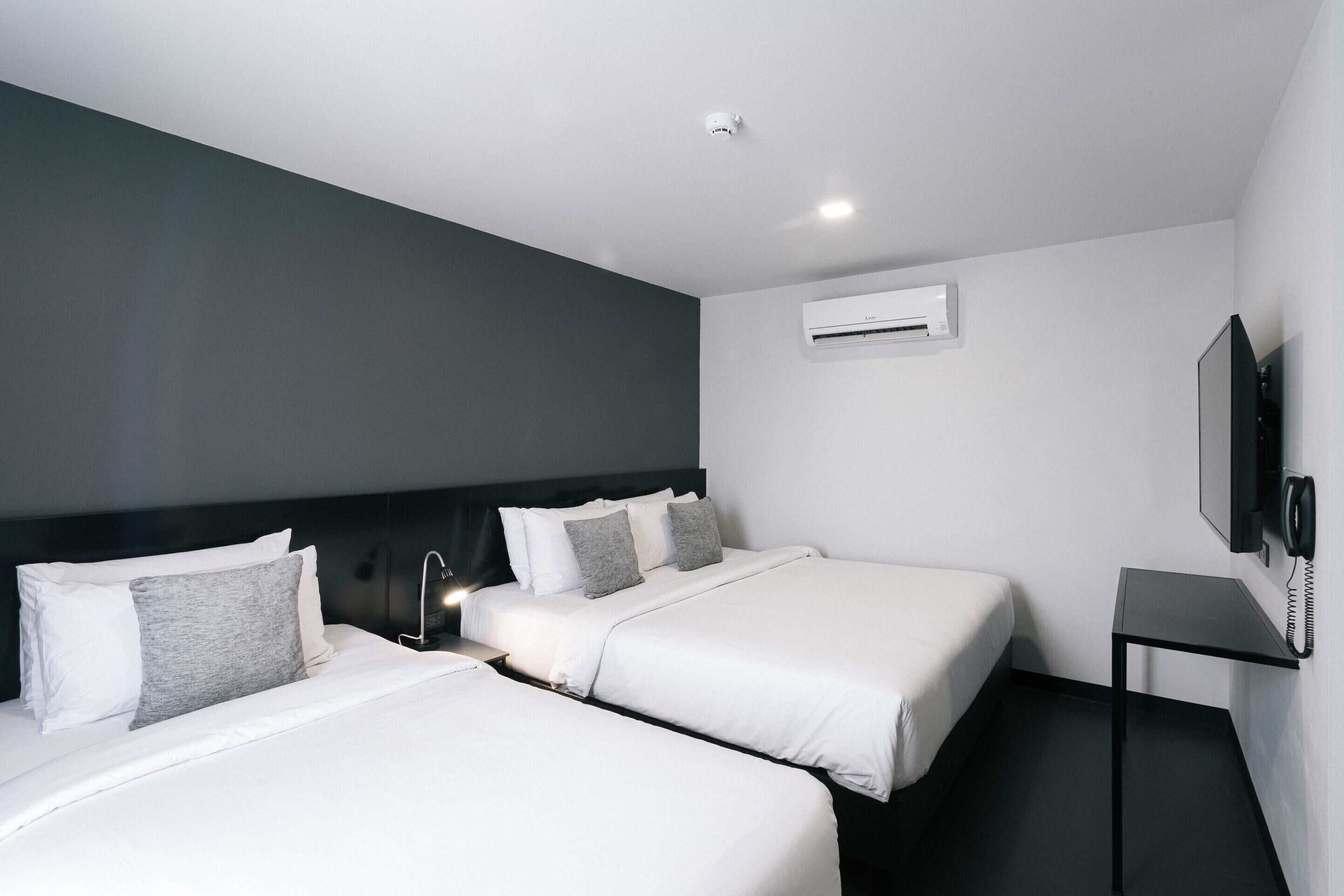 A hotel room with two beds, a flat-screen TV, a bedside table, and an air conditioning unit.