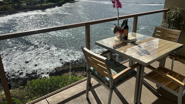 A balcony with a table and chairs overlooking a coastal view with palm trees and ocean waves.