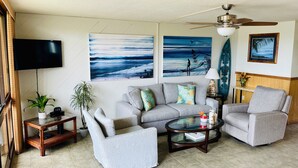 A living room with a grey sofa, two armchairs, a coffee table, a TV, and artwork depicting ocean scenes.