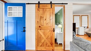 Een kamer met een blauwe deur, een houten schuifdeur in de stijl van een schuur, een witte nachtkastje met een lamp, een grijze bank en een raam met jaloezieën.