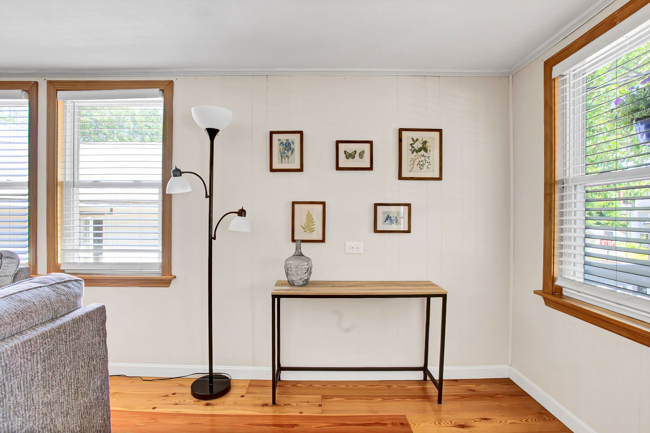 A room with a wooden floor, a grey sofa, a black floor lamp, a small table with a vase, and framed pictures on the wall.