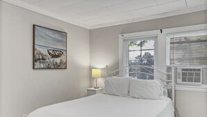 Een slaapkamer met een bed, nachtkastje met een lamp, raam met zonwering en een schilderij aan de muur.