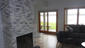 Una sala de estar con chimenea de piedra, un sofá, un sillón y vista a una masa de agua a través de ventanas amplias.