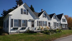A two-story white house with black shutters, a front porch with Adirondack chairs, and a chimney. - Remarkable Four Gable House Overlooking Friendship Harbor (Friendship)