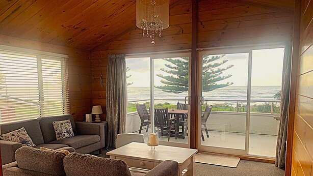 A cozy room with a sofa, a coffee table, and a dining table with chairs. There is a large window with a view of trees and a body of water. - Ohope Holiday Apartment (Ohope)