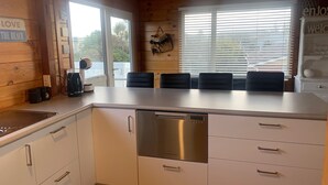 A modern kitchen with white cabinets, a stainless steel dishwasher, and a large window with blinds. - Ohope Holiday Apartment (Ohope)