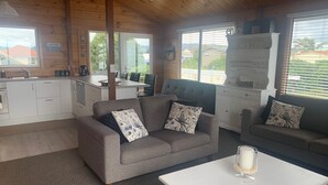 A cozy cabin-style room with a grey sofa, a white table, and a kitchen area. - Ohope Holiday Apartment (Ohope)