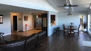 A spacious living area with a dark wood floor, a leather couch, a dining table with chairs, and a kitchen area with a refrigerator and bar stools. - Large Lake House 30 Feet From The Water With Private Dock Out The Door.  (Mears)