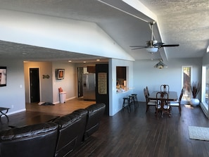 A spacious living area with a dark wood floor, a leather couch, a dining table with chairs, and a kitchen area with a refrigerator and bar stools.