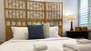Een net opgemaakt bed met witte lakens, een blauw geruit kussen en twee opgerolde handdoeken.