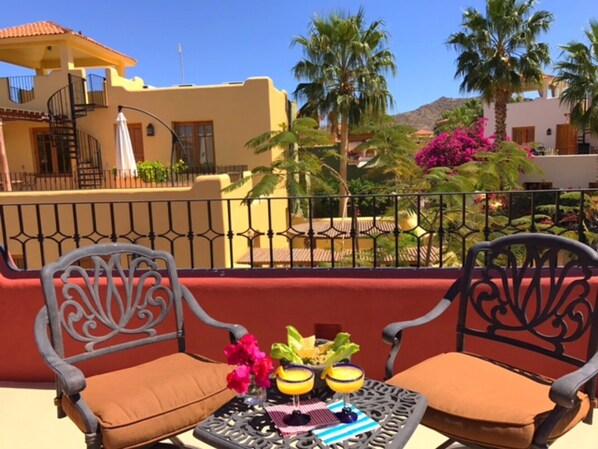 A balcony with two metal chairs, a small table with a drink and a salad, and a view of a yellow building with palm trees and a mountain in the background.