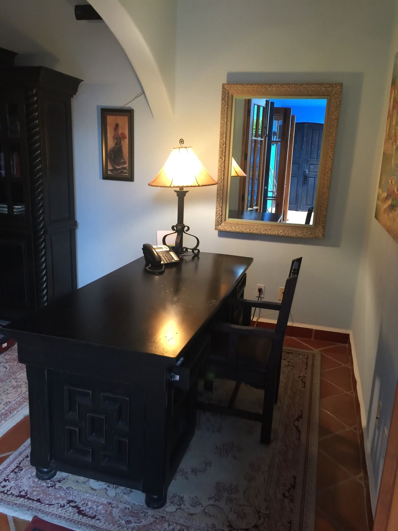 A dining area with a table, chairs, a lamp, a telephone, a framed mirror, and a painting on the wall.