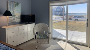 A room with a white dresser, a television, a wicker chair, and a large sliding glass door leading to a deck with a view of a house and a car. - Bright & Airy 3BR Home with Views of Lewis Bay! (1498) (West Yarmouth)
