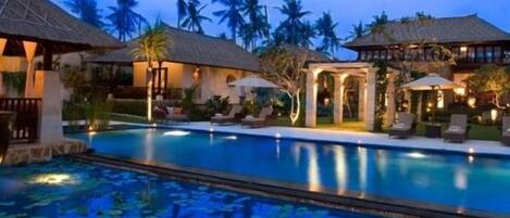 Zona de piscina con sillones de descanso y sombrillas, rodeada de edificios con techos de paja y palmeras.