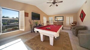 A pool room with a pool table, a ceiling fan, and a large window overlooking a landscaped area.