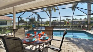 A covered outdoor area with a pool, dining table, and chairs, overlooking a body of water and palm trees.