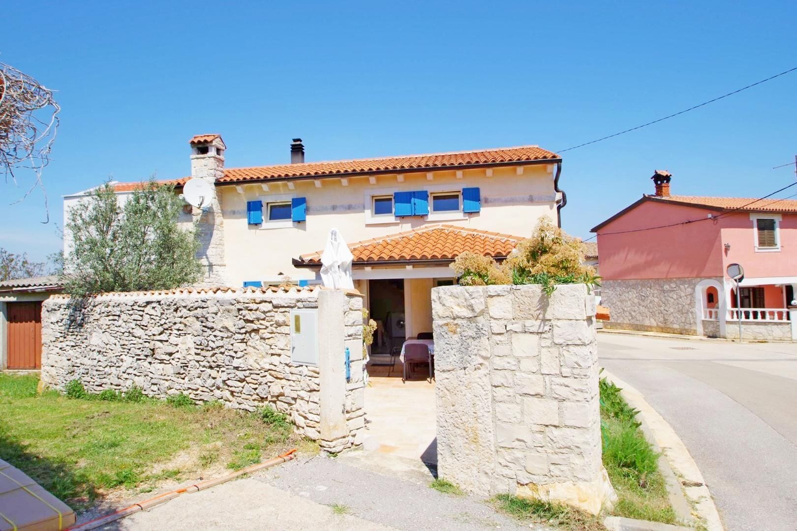 A two-story house with a stone wall entrance, a tiled roof, and blue shutters.