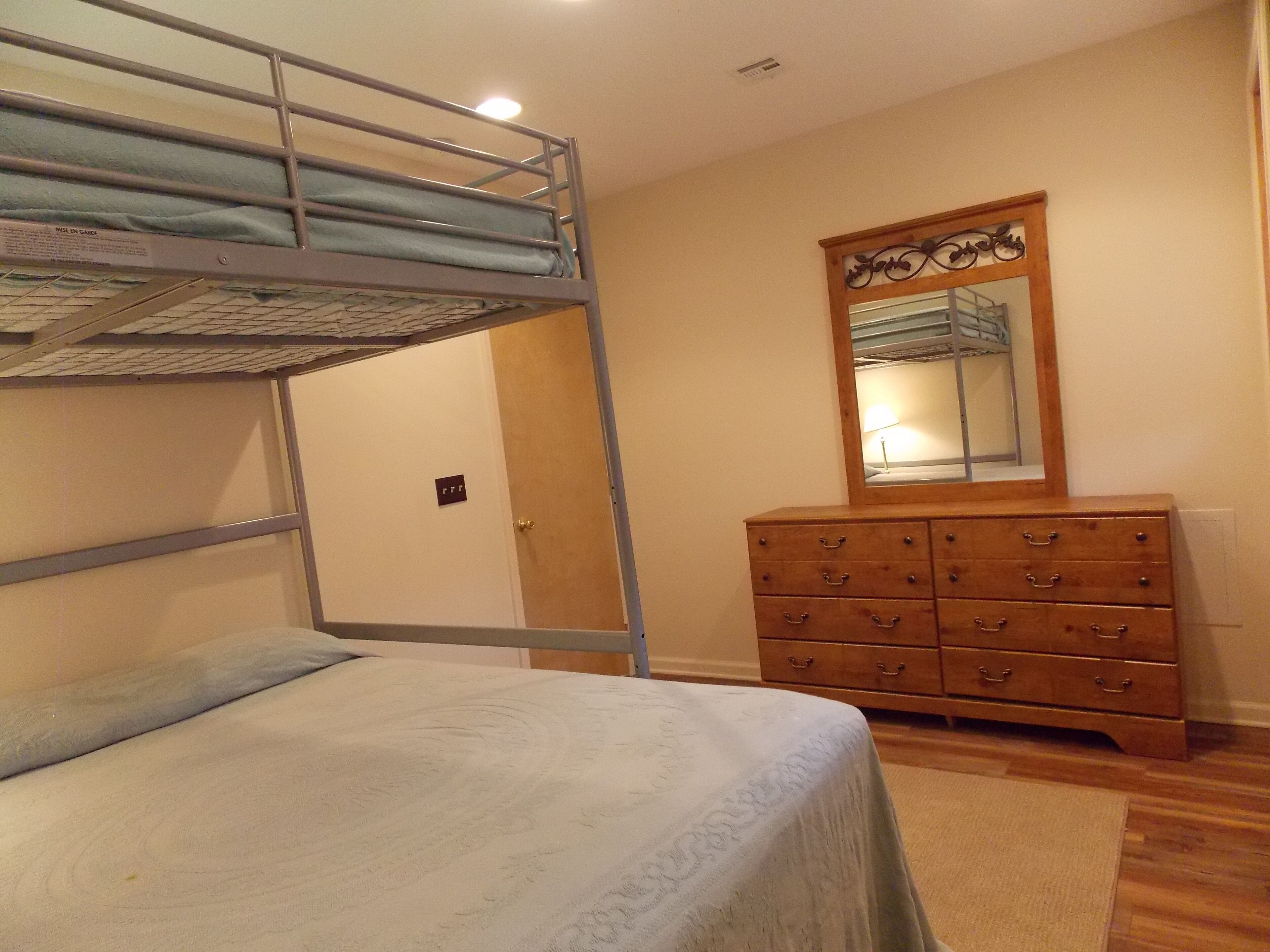 Una cama litera con colchón, un aparador de madera con cajones, un espejo y una puerta con un letrero.