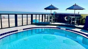 A swimming pool with a tiled edge, a lifeguard stand, and two umbrellas. - OF Condo Gone2TheBeach @ Symphony Beach Club (Ormond Beach)