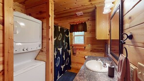 Baño de una cabaña de madera pequeña, con lavadora, lavamanos y espejo.