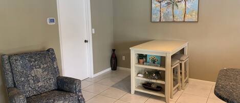 A living room with a patterned armchair, a small white table with decorative items, and a painting of palm trees on the wall.