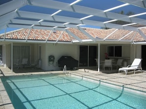 Ein Schwimmbecken mit Fliesen umgeben, eine überdachte Terrasse mit Stühlen und ein Grill.