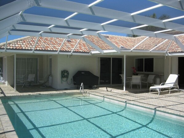 Una piscina con deck de baldosas, un patio cubierto con sillas y una parrilla.