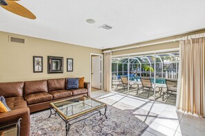 A living room with a brown leather sofa, a glass coffee table, and a patterned rug. There is a glass door leading to a patio with outdoor furniture and a pool.