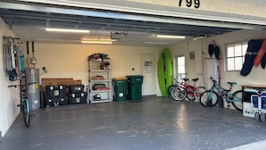 Un garage avec le numéro 799 peint sur le mur, un chauffe-eau, des étagères remplies d’outils et d’équipements, une planche de surf et des vélos.
