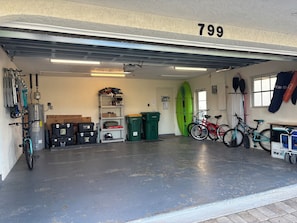 A garage with a number 799 on the wall, a water heater, shelves with tools and equipment, a surfboard, and bicycles.