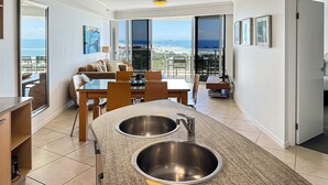 Una cocina moderna con dos lavamanos de acero inoxidable, una zona de comedor con mesa y sillas, y vista al océano a través de amplios ventanales.