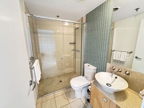 A modern bathroom with a glass-enclosed shower, a white toilet, a sink with a granite countertop, and a mirror with a towel rack. - Resort Apartment in Maroochydore (Maroochydore)