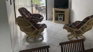 En stue med to fletstole, et fjernsyn på et stativ, et bord med en glasvase og en dør, der fører ud til en balkon.