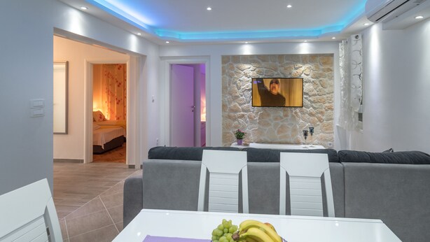 A modern living room with a stone wall, a flat-screen TV, grey sofas, and a dining table with fruit.