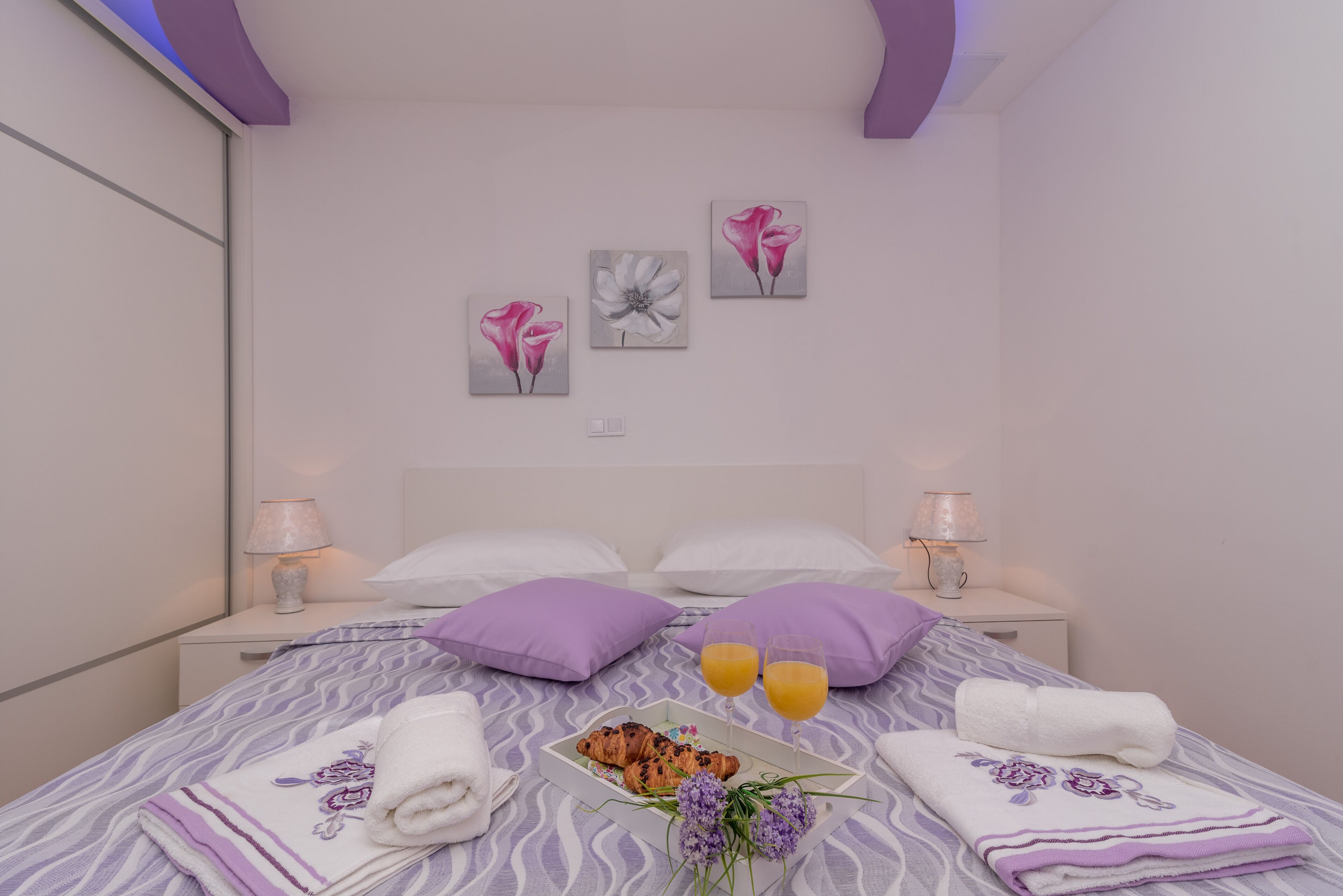 A neatly made bed with pillows, a tray of croissants, two glasses of orange juice, and towels.